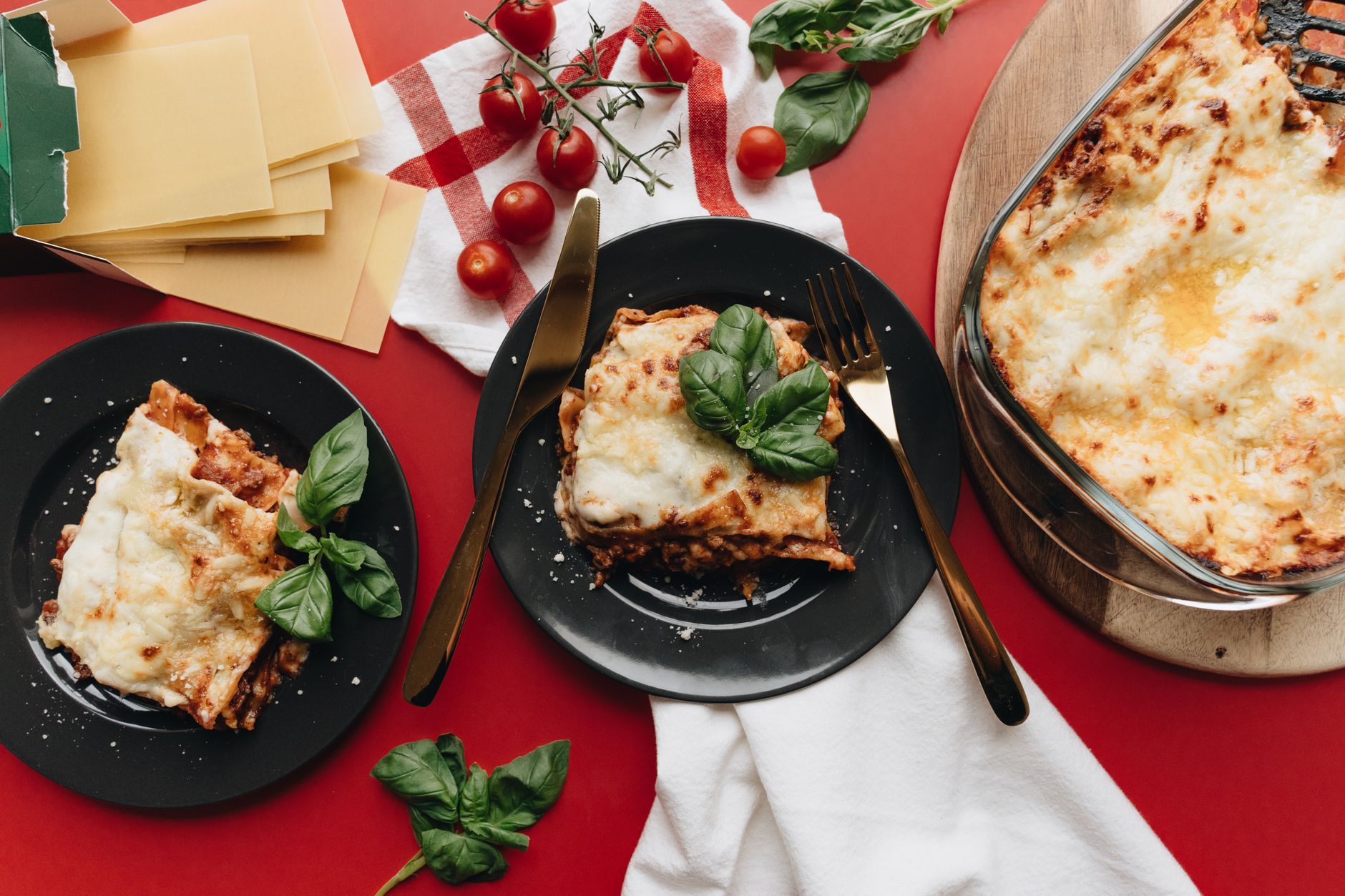 Lasagnes gratinées dans un plat en verre et en parts dans des assiettes noires, avec basilic, tomates cerises et feuilles de pâte, sur fond rouge.