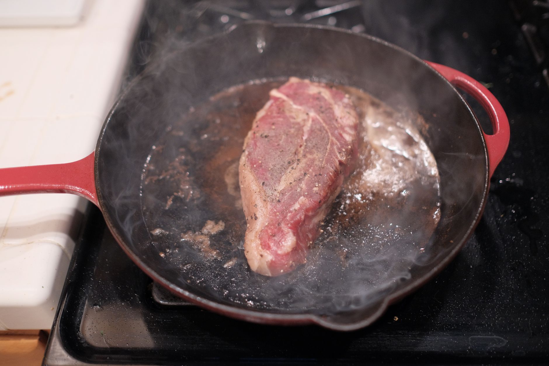Poêle en fonte rouge sur une cuisinière, avec une pièce de viande poivrée en cours de cuisson et de la fumée.